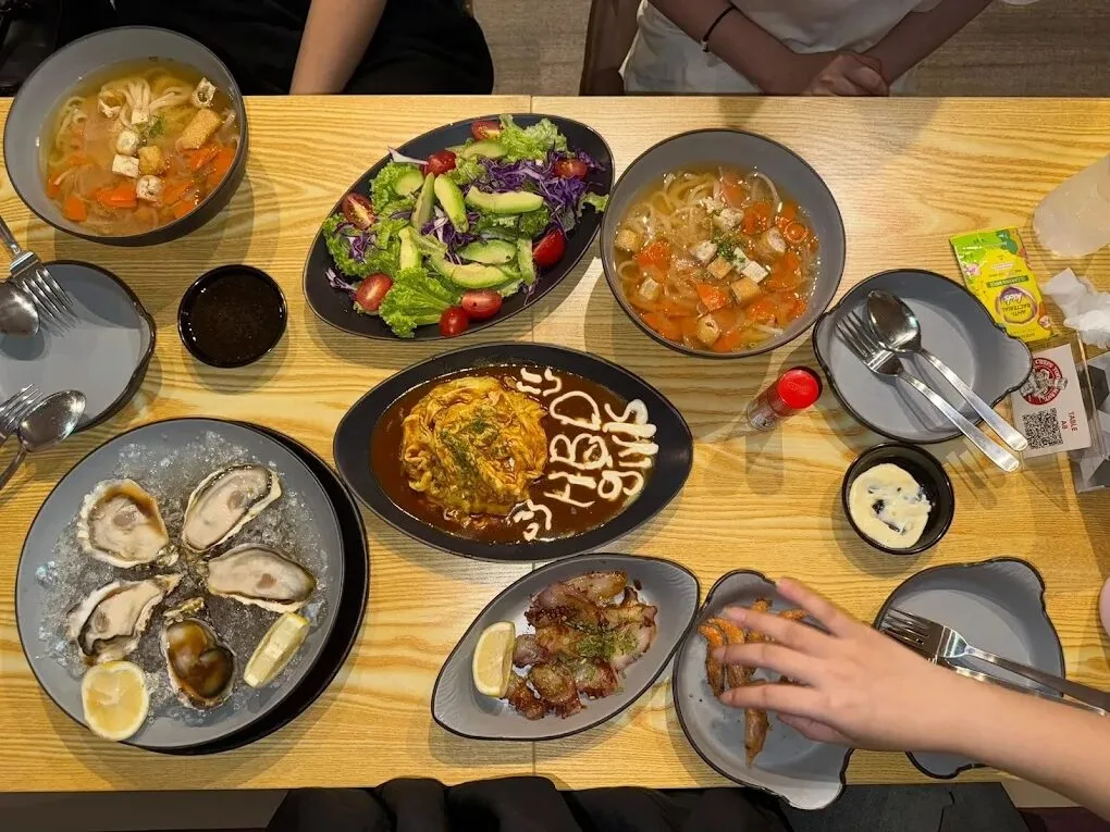 A table spread at 2 Chefs Too Many — omu rice, udon, oysters, salad and skewers on wooden tables